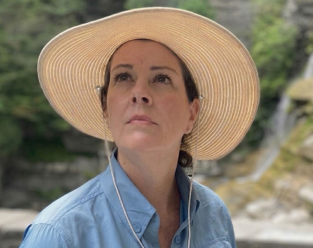Heather, owner of Nest Well Professional Organizing, looking upward in a natural outdoor setting, wearing a wide-brimmed hat and blue shirt