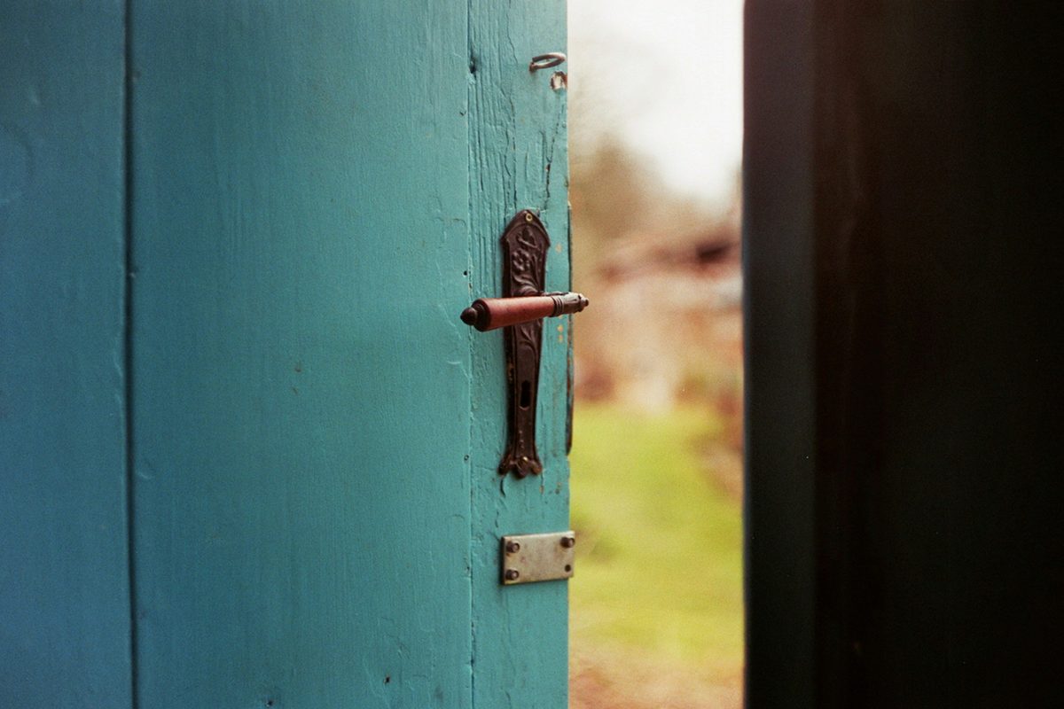A teal blue door standing slightly ajar, vintage iron handle, with a glimpse of a green garden beyond